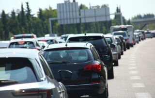 A long line of cars sits in heavy traffic on a multi-lane highway during a bright, sunny day, with a large overhead road sign in the background.
