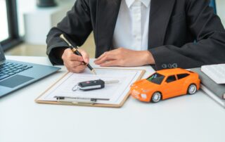 A person wearing a black blazer and white collared shirt sits at a white desk, holding a black and gold pen to sign a document on a clipboard. On the clipboard rests a car key fob, and to the right sits a bright orange toy car model. A laptop is visible on the left side of the desk, and a calculator sits on top of notebooks to the right.