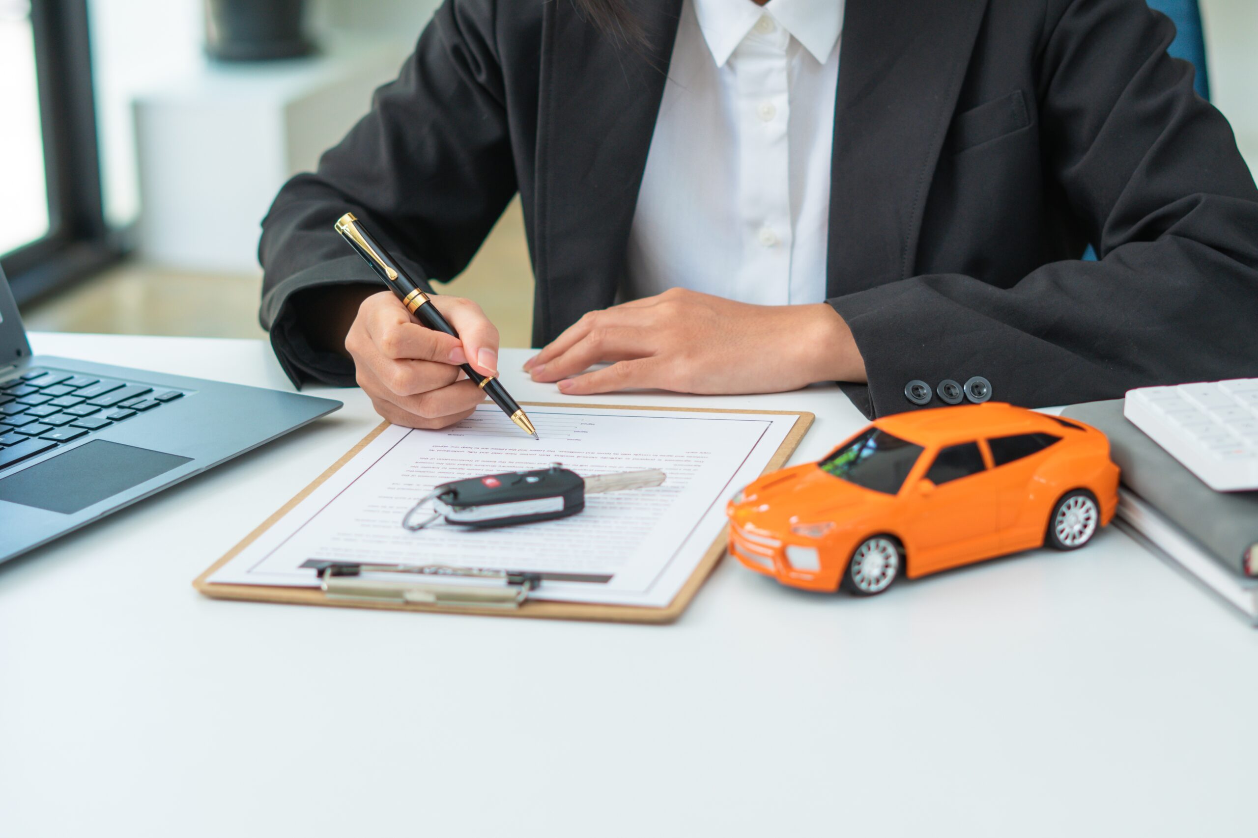 A person wearing a black blazer and white collared shirt sits at a white desk, holding a black and gold pen to sign a document on a clipboard. On the clipboard rests a car key fob, and to the right sits a bright orange toy car model. A laptop is visible on the left side of the desk, and a calculator sits on top of notebooks to the right.