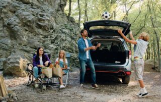 A family of four enjoy an afternoon of camping outdoors. The mother sits on a folding chair on the left, next to a campfire structure and a table holding groceries. A young girl sits next to her, looking toward her father and brother. The father stands behind the vehicle, throwing a soccer ball into the air, while his son stands opposite him, arms raised to catch it, next to the open trunk of a silver SUV. The family is surrounded by a dense forest, with a large rock formation rising behind the mother and daughter.