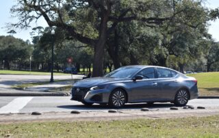 A dark gray Nissan Altima sedan drives along a paved road or intersection in a park-like setting. Large, mature oak trees with spreading branches provide shade in the background, next to a grassy lawn area. A street lamp, a yield sign, and other distant parked cars are visible under a clear, bright blue sky.