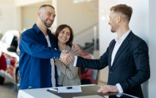 An alt text for this image could be: A smiling couple stands at a dealership counter, shaking hands with a car salesperson dressed in a dark suit jacket and white collared shirt. The man, wearing a blue button-down shirt over a white T-shirt, has his arm around the woman, who wears a patterned blazer and smiles warmly at the salesperson. A car key and a document lie on the white counter between them, while a red car is softly blurred in the background of the bright showroom.
