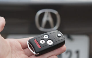Hand holding a black Acura key fob remote in front of the Acura logo on a car trunk.