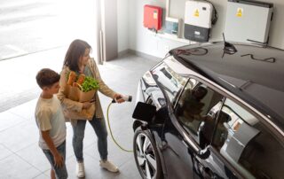 Woman holding groceries and plugging a charging cable into a black electric car in a home garage alongside a young boy.