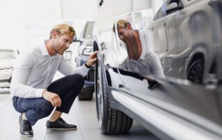 "A man in a white dress shirt crouching down to inspect the front wheel and tire of a car in a showroom.