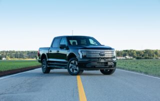 A black Ford F-150 Lightning electric pickup truck parked in the center of an empty paved road surrounded by fields.