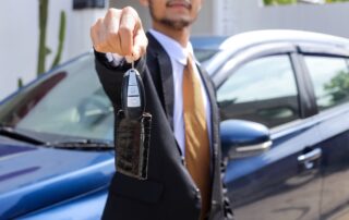 A car salesman in a suit handing over car keys in front of a blue vehicle.